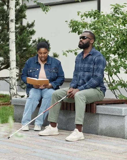 Disabled blind man sitting outside with carer while she reads to him